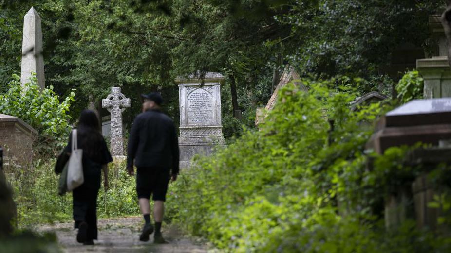  Two visitors walk through Highgate Cemetery. In the foreground is green bushes, in the background are headstones