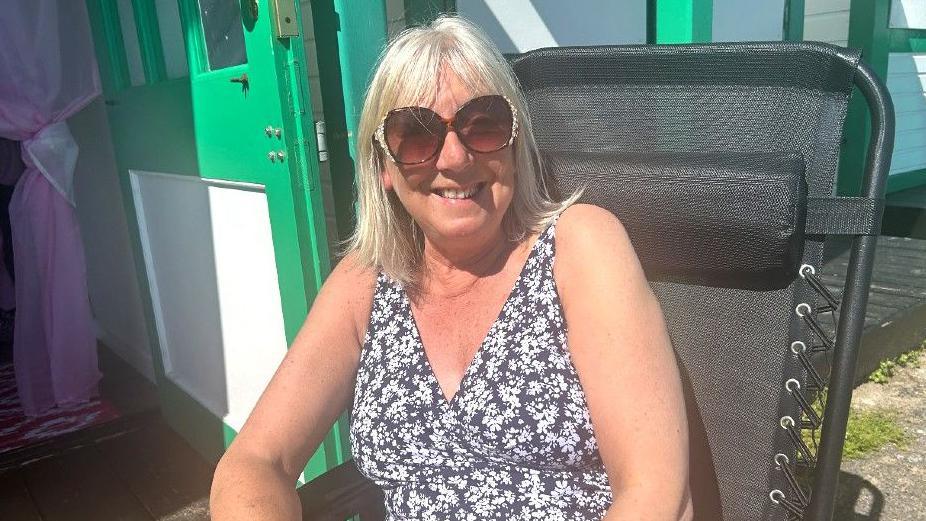 Liz Symons sits on a black, collapsible garden chair on the path outside her beach hut. She has very light-blonde bleached hair which is shoulder-length with a fringe. She is smiling, lightly-tanned, wears large sunglasses and a v-neck either sleeveless top or swimming costume which is purple-blue with a pattern of small white flowers. To her left one door of the hut is open showing a pink voile, see-through curtain which has been tied back in a knot half way down.