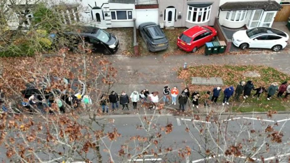 A long line of people shown from above. They are stood side by side and can be seen through the brown leaves of some trees. 