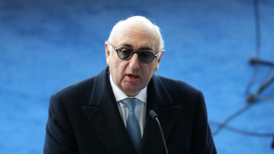 Barnett with thinning white hair, wearing darkened glasses, a dark overcoat, white shirt and blue tie, stands in front of a microphone in 2018.