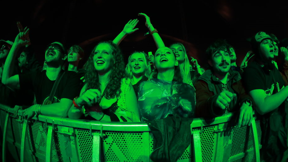 A crowd of young men and women smile and raise their arms as they look towards a stage, shrouded in a green light