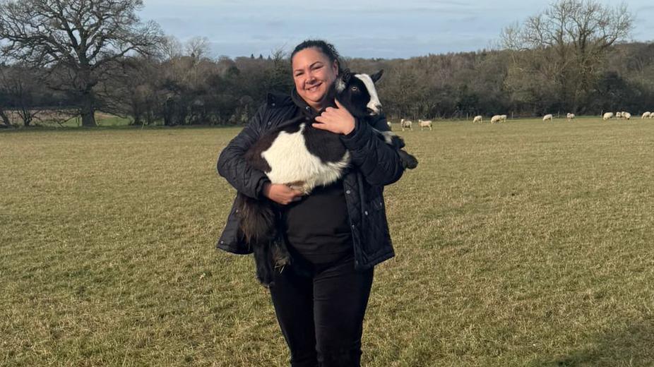 Olivia Mikhail holding Comet a black and white goat in her arms, in a field, with sheep behind her. She is wearing dark trousers, a coat, and boots, is smiling and looking straight at the camera. 