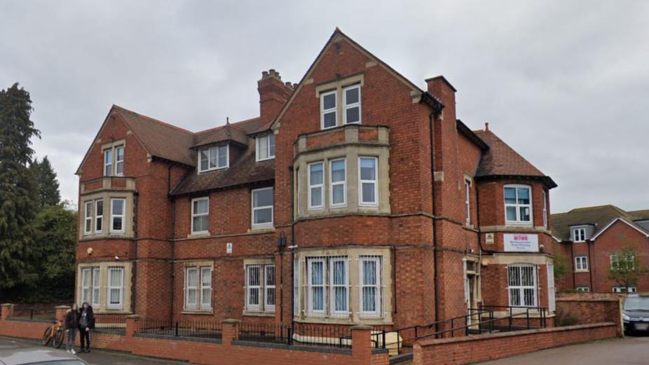 Large red bricked three-storey building with four sets of bay windows. It has black railings around the perimeter of the building. 