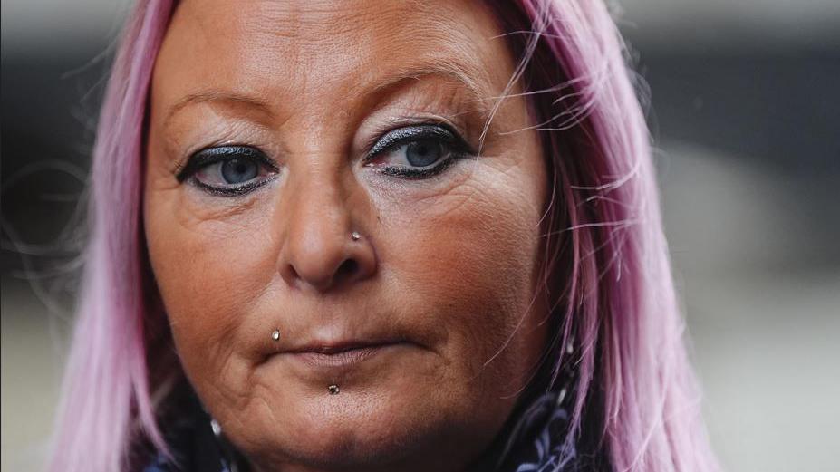 Woman with a tan and purple tinted hair. She appears solemn and is looking off camera.