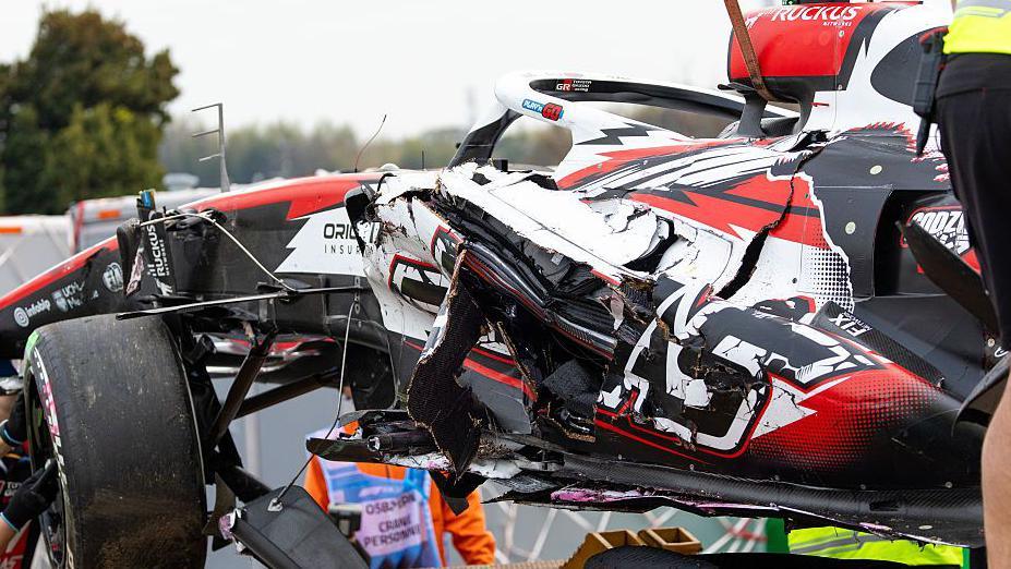 A red, white and black F1 car being lifted up onto a trailer after a crash. The body work on the car is significantly damaged. Workers are helping to guide the car onto the trailer.