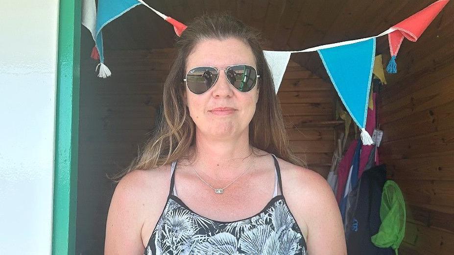 A head and shoulders shot of Sally Thomas, who is standing in the doorframe of her beach hut. She had long brown hair with lightened ends, is wearing wire-rimmed sunglasses and is smiling slightly. She wears a spaghetti-strap black and white top with a pattern of fronds on it, and what looks like straps from a swimming costume underneath. She also has a thin silver chain with four or five rings looped on at the front of it. Immediately behind her head is turquoise, white and red bunting. Items for the beach can be seen on one side of the cabin in the shade.