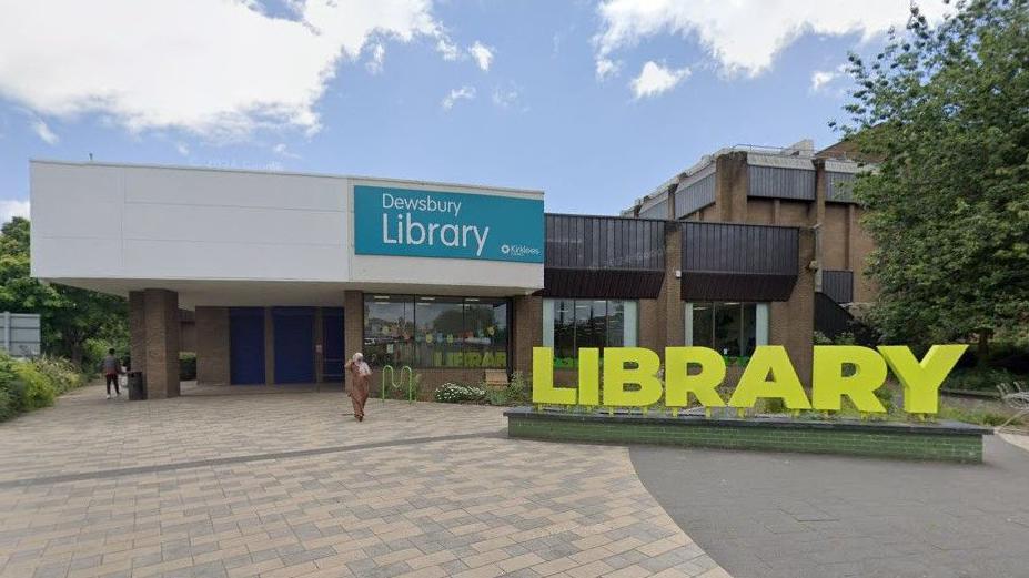 Dewsbury Library in its current location - a single storey building with a large yellow sign outside which reads "library"