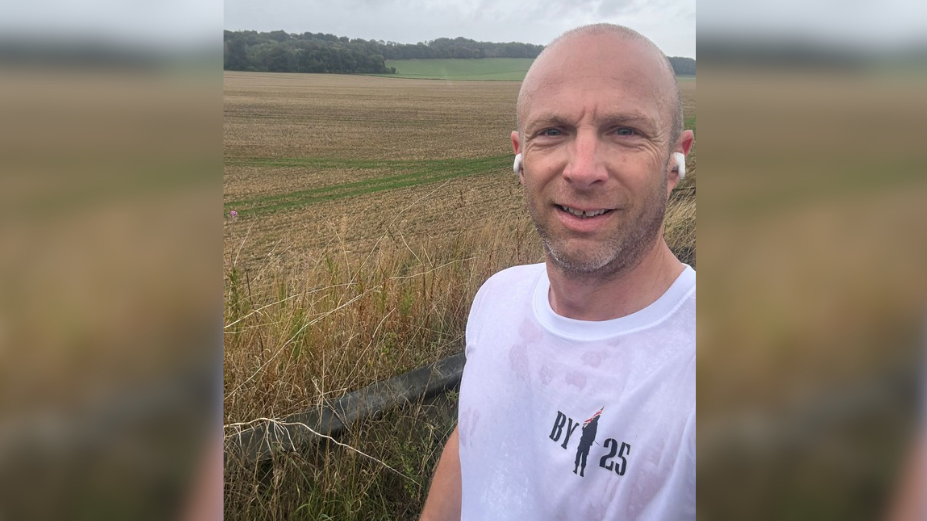 Man in white t-shirt which says 'BY 25'. He has ear pods in his ears. There is a field in the background.