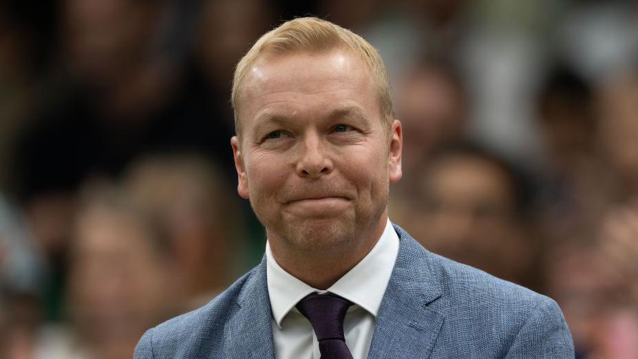 Sir Chris Hoy smiles shyly as the crowd give their appreciation at Wimbledon. He is wearing a grey suit, white shirt and black tie and the spectators are out of focus in the background.