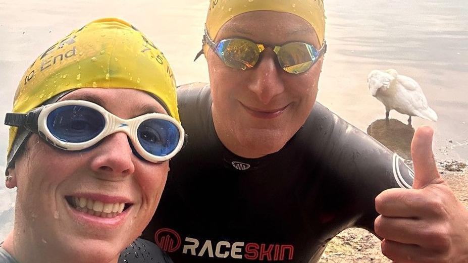 A smiling woman and man stood next to each other with water in the background. They are both wearing wetsuits, swimming goggles and yellow swimming hats. The man is posing with his thumb up.