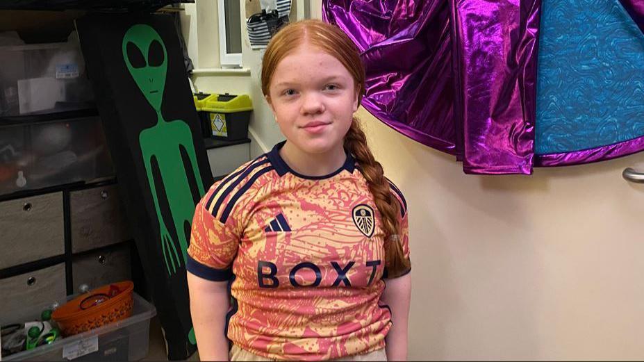 Jessica looks at the camera. She is wearing a football shirt. A green alien painted onto a board is propped up behind her and a Halloween costume is hanging up next to it.