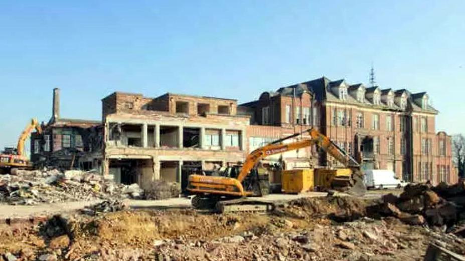 A yellow bulldozer digs through rubble in front of part-demolished, red-brick hospital buildings at Lincoln County Hospital.