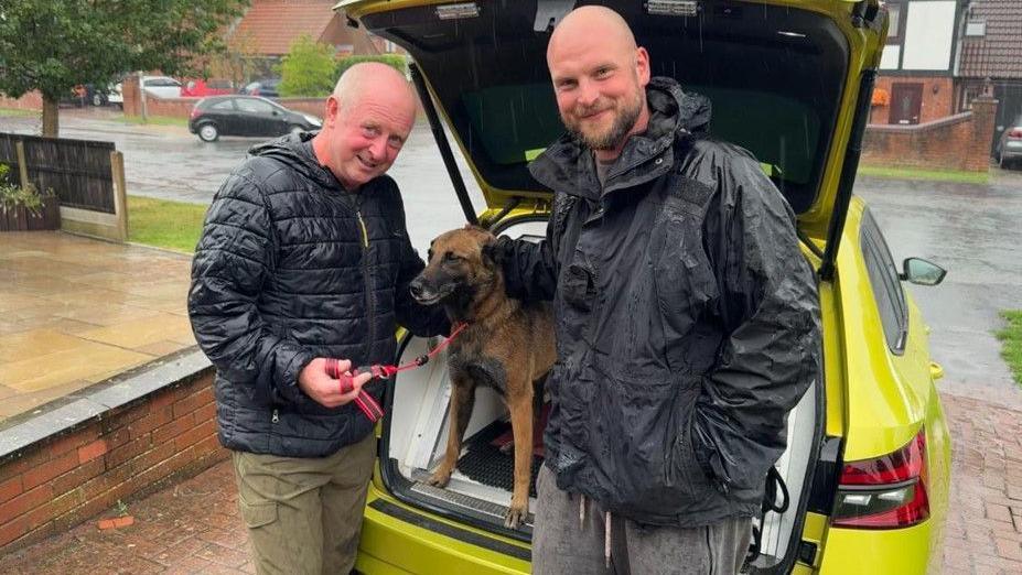 PC Tune stands to the left of the picture and Phil is on the right. It is raining and they both wear black jackets. The boot of a bright green/yellow car is up and Quinta stands in the back. PC Tune holds her red lead.