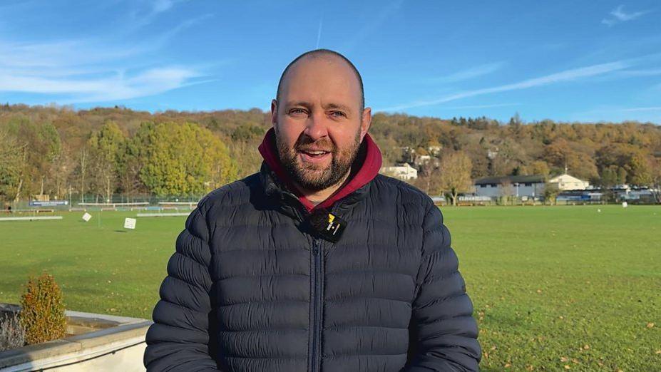 Rory wearing a dark puffer jacket with a red hood underneath stands outdoors on a sunny day. Behind him is a large grassy field bordered by a low concrete wall with small shrubs. In the background, there are trees with autumn foliage and a few houses scattered along a hillside under a clear blue sky.
