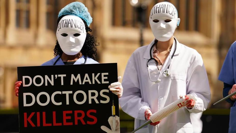 Two people dressed as doctors, one carrying a sign reading 'don't make doctors killers'