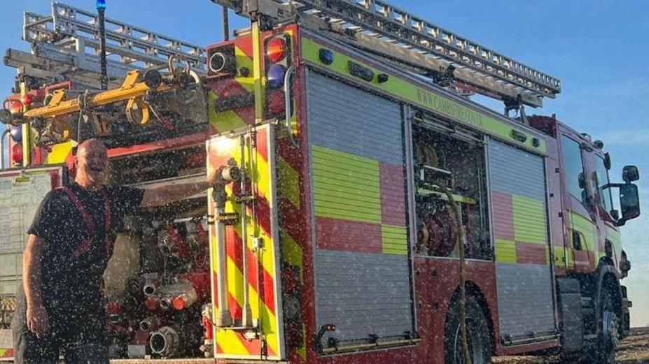 A grainy photo of Mervyn Housden wearing a blue t-shirt and dark firefighter trousers with red braces standing in front of a fire engine and laughing