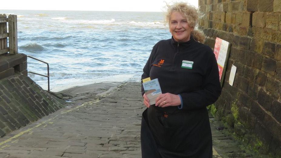 A woman stands by the sea and holds up a passport.