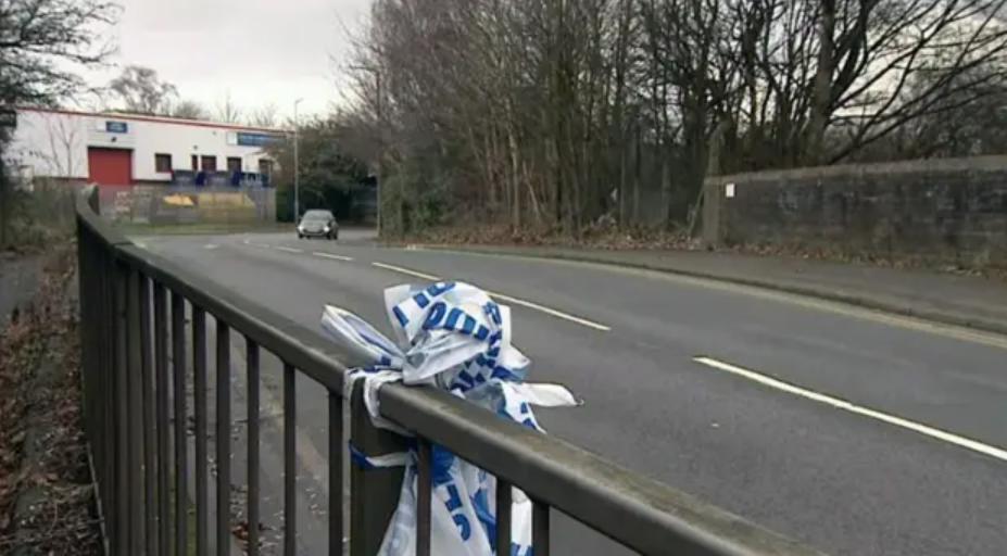 A metal fence beside a road with blue and white police tape knotted around it and trees on the other side