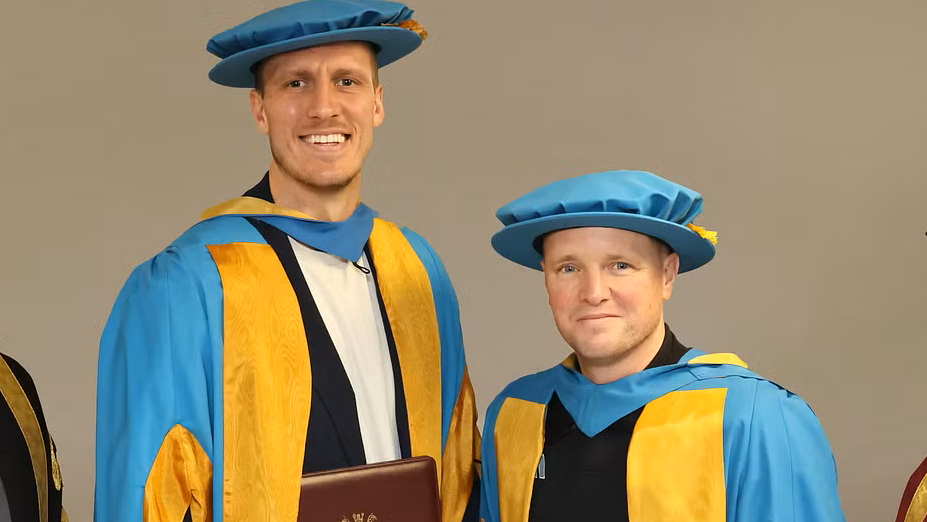 Dan Burn and Eddie Howe smiling into the camera. They are both wearing bright blue hats and matching robes with golden trim. Burn is on the left and has light hair and stubble and is wearing white underneath the robe. He is considerably taller than Howe. Howe is smiling with his mouth closed and has black material peeking out of the colour of the robe. His hair is mostly covered by the hat.