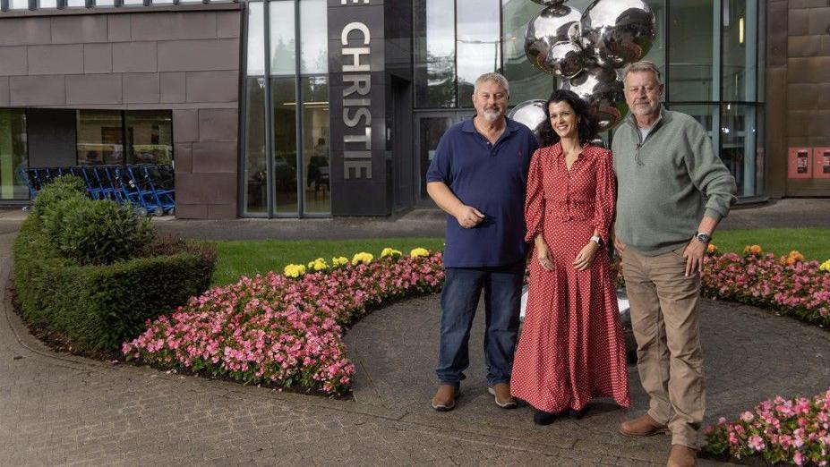 Glenn and Russell Wood stand outside the entrance to The Christie, with Dr Serra standing in between them. They are all smiling. Dr Serra has long brown hair and is wearing a long red dress.
