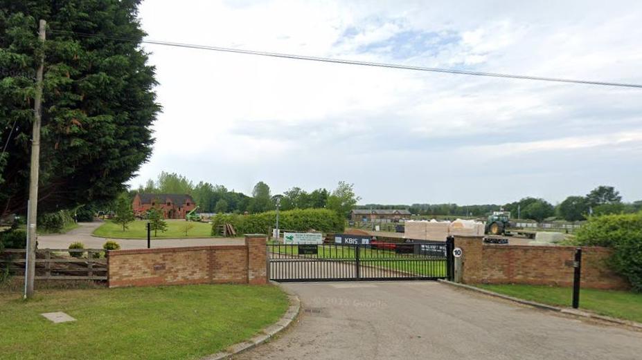 Entrance to the equestrian centre showing wide black metal gates with brick walls on either side. There is a gate and a tree to the left. There are fields behind the gate and some low buildings. There are some clouds in the sky.