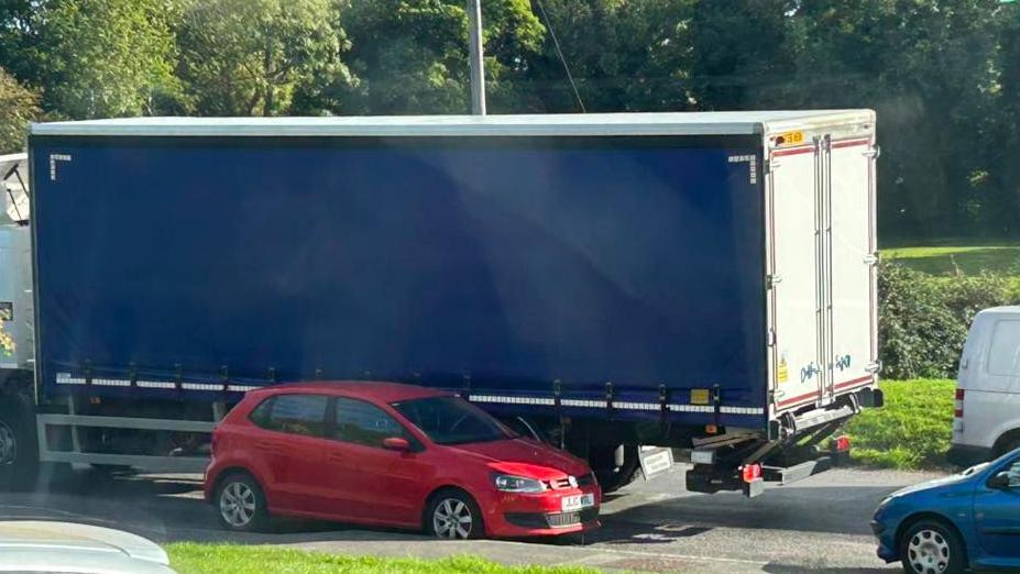 A large blue lorry passes a red parked car with trees and grass in the background of the image.