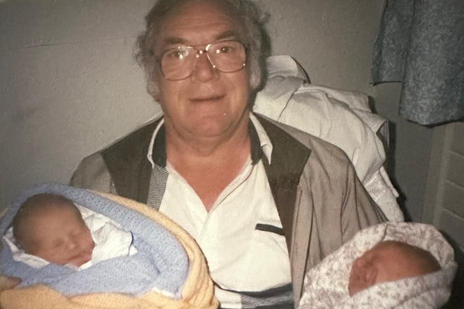 Brian Teasdale, in his 60s and wearing a white shirt, grey jacket and glasses. He is holding twin babies wrapped in blankets.