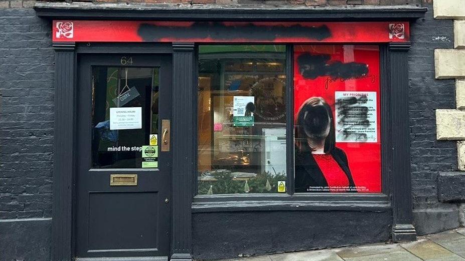 A black shop front has black spray paint on its windows and red shop sign. There is spray paint covering a window decal of Julia Buckley's face.