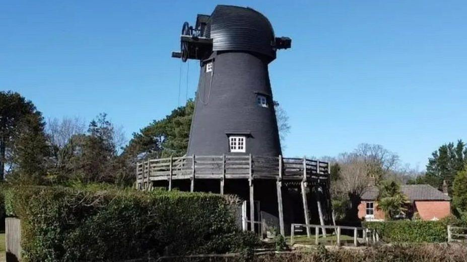 Bursledon Windmill, pictured near greenery and with a house in the background. It has wooden fencing around it and the windmill is painted black.