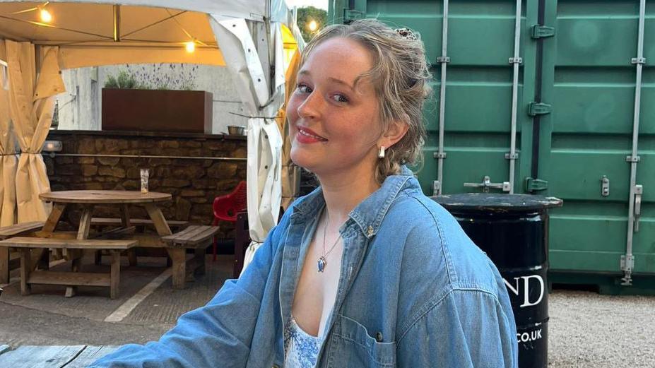 Mollie Mulheron wearing a denim jacket with a blue necklace and her blonde hair tied up, smiling at the camera