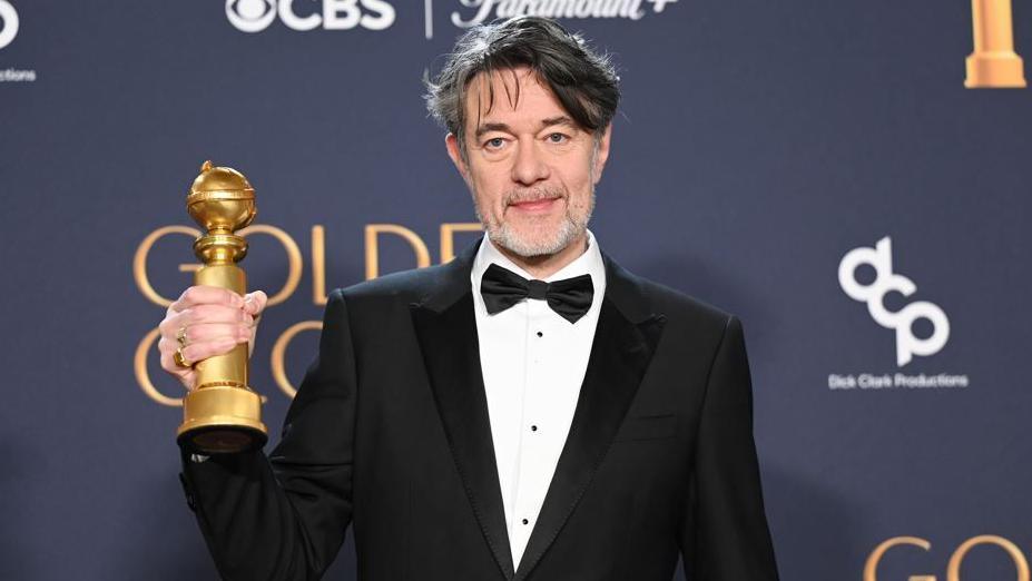 Peter Straughan, wearing a tuxedo, stands in front of an advertising board,  lifting his Golden Globe award with his right hand after winning best Adapted Screenplay in January 2025. 