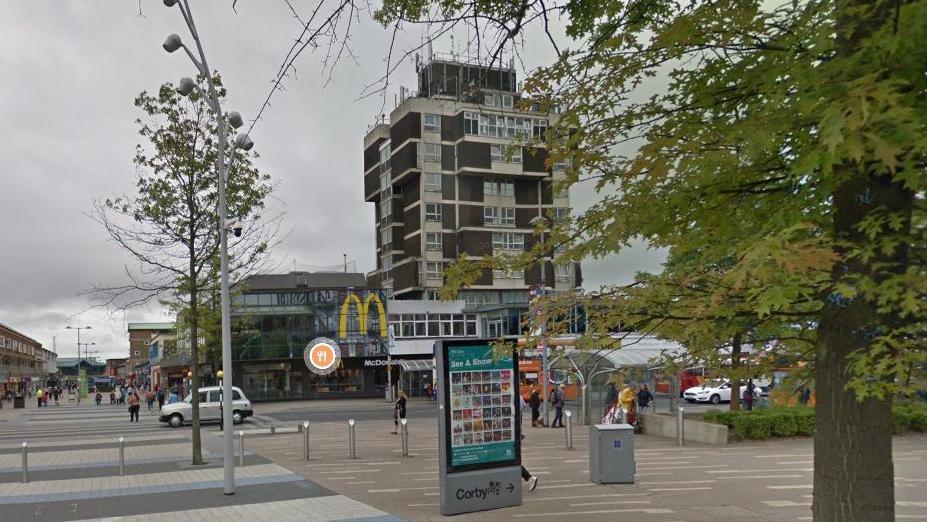 1980s eight-storey office block in red brick with white lines around each floor and white window frames. The bottom floor is occupied by shops and there is a large letter M in yellow to the right of the eight storeys. There is a small tree to the right of the building and a larger tree in the foreground to the left. There is a road passing the office building and a dynamic advertising board is visible in the centre of the picture.