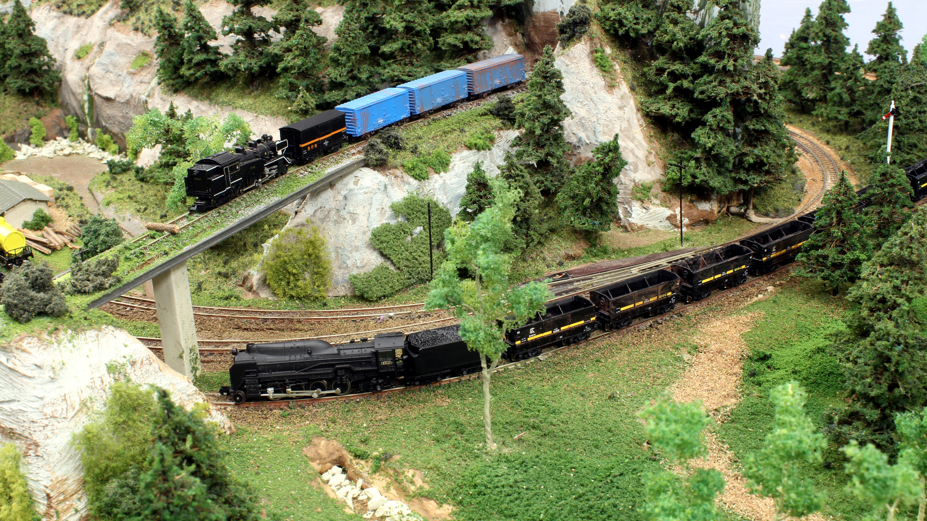 A model railway exhibition showing a rural scene with one train passing under a bridge as another train crosses under it. 