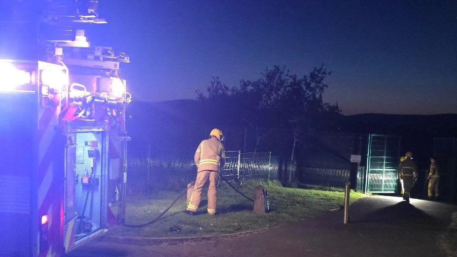 A number of firefighters stand near to a fire engine with its lights flashing.
A hose can be seen being brought towards the gates of the play park.
