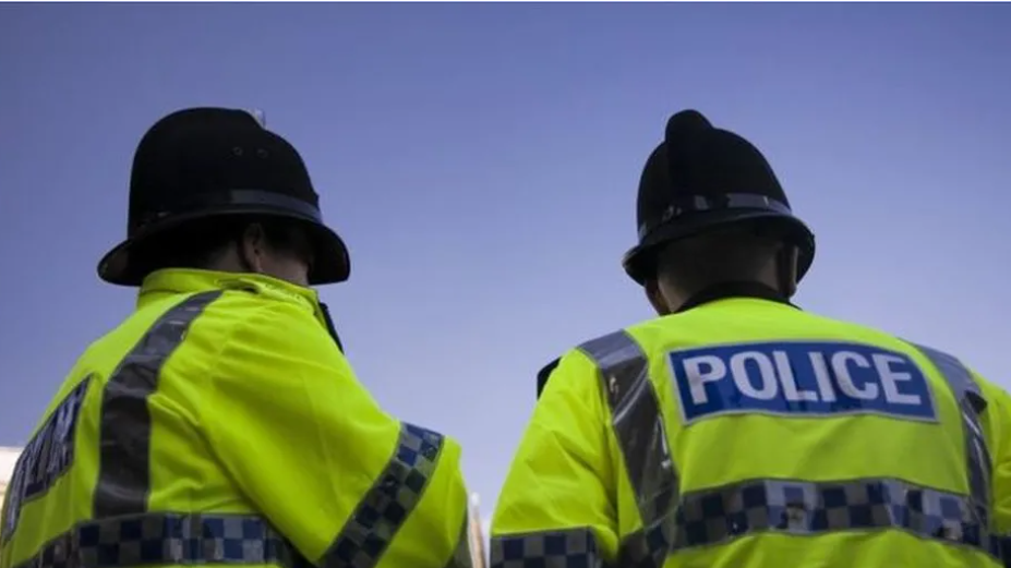 Two British police officers in black hats and bright yellow jackets with the word "police" in white lettering on a blue rectangle background. 