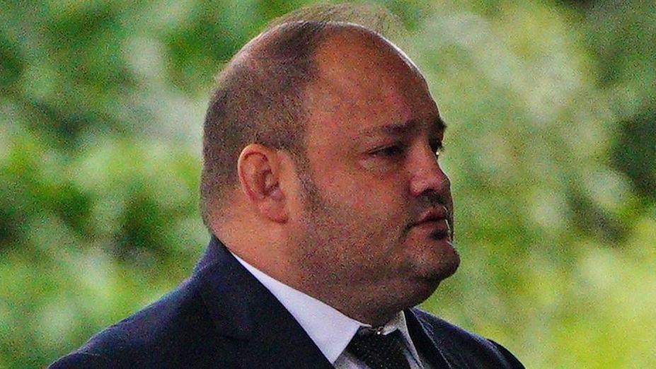 John Stringer outside court in front of greenery in the far background. He is a heavy set man, bald on top with short cropped hair on the sides and with stubble