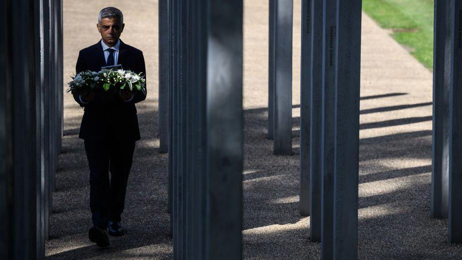 Sadiq Khan carries a wreath through the 7/7 memorial, which consists of 52 vertical stainless steel columns. He is wearing a dark suit and tie and white shirt. 