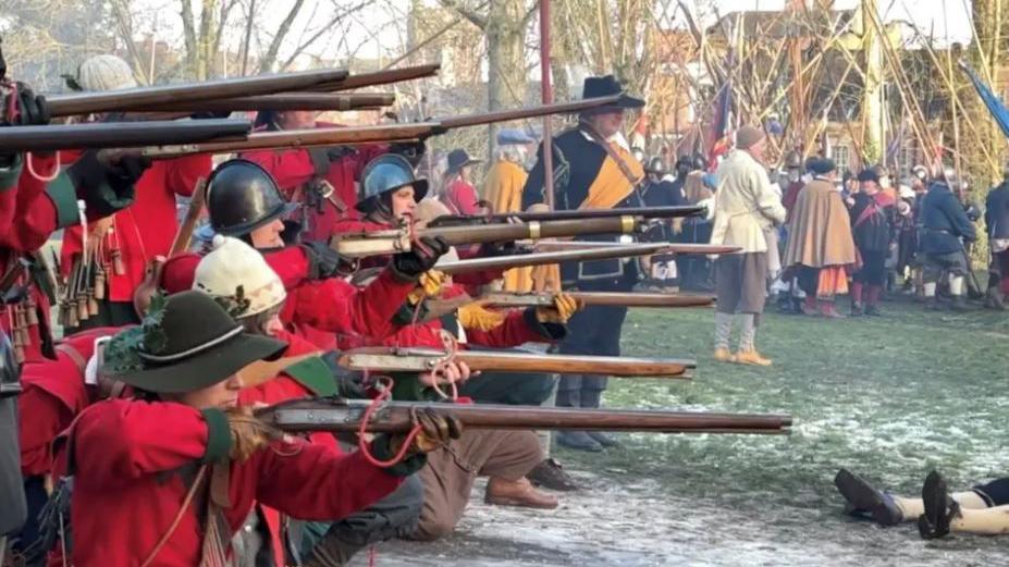 A group of people stand holding replicas of guns and weapons in a field in a reenactment of the 1644 Battle of Nantwich.