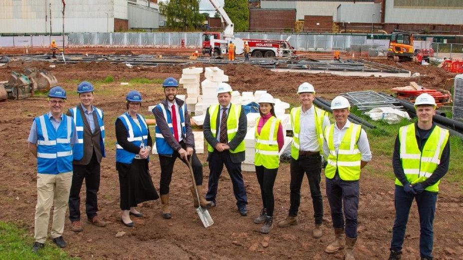 Staff from Wye Valley NHS Trust and contractor Speller Metcalfe at the site of the new community diagnostic centre in Hereford. They are wearing high-viz vests and hard hats, and are standing on rough ground. One of the men is holding a spade.