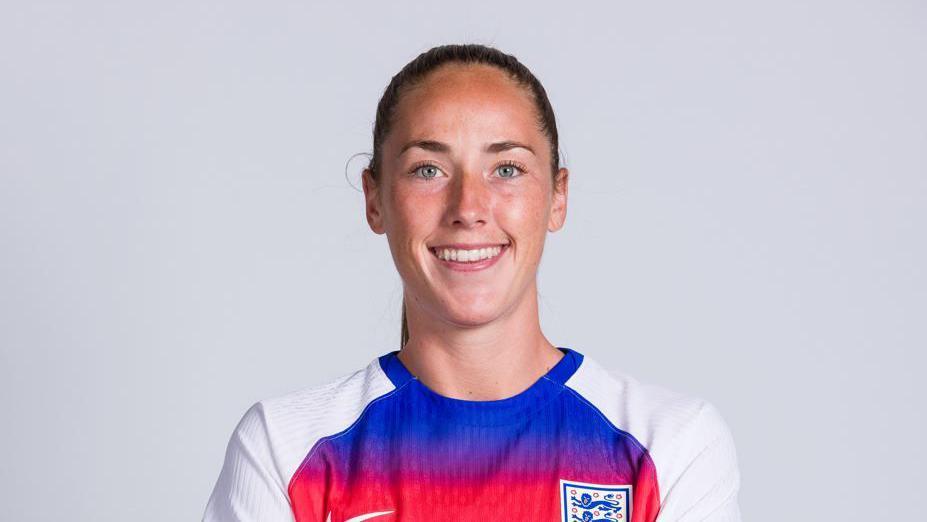 A woman with tied back brown hair in a white shirt with a blue and red pattern on the front