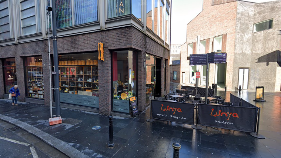 Exterior view of Lunya restaurant on the corner of Hanover Street and College Lane in Liverpool. The restaurant is a grey brick building with a small amount of outdoor seating enclosed by canvas netting bearing the company's name.