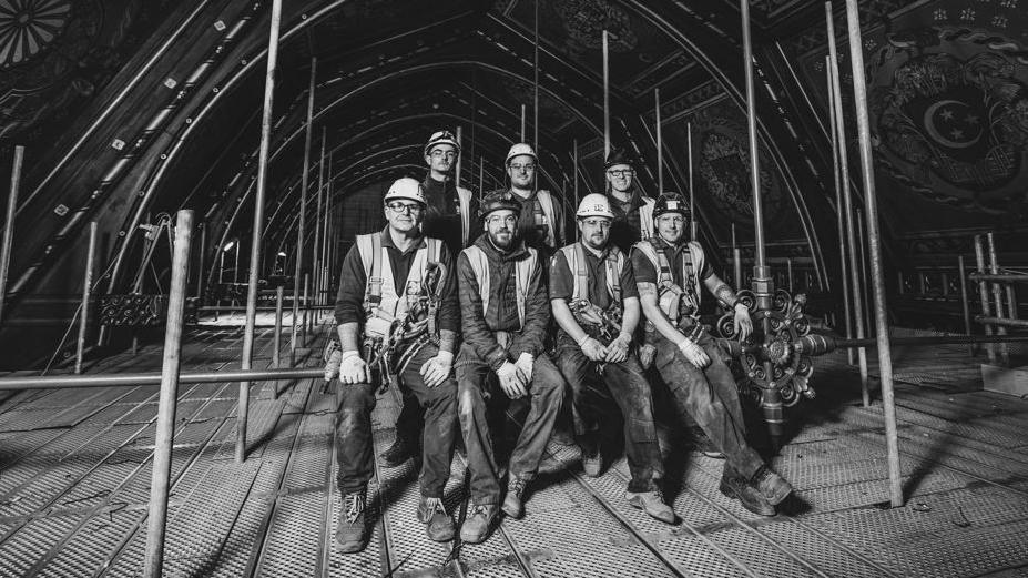 A group of tradespeople in safety gear including hard hats pose for a photo on a scaffold underneath the town hall roof. 