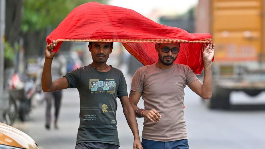 The image shows two men using a red scarf to protect themselves from the heatwave during a hot summer afternoon in Delhi in 2024. 