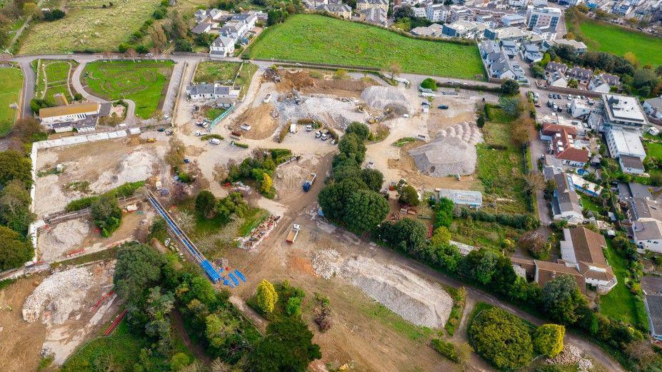 Aerial shot of demolition site at Overdale in Jersey. Green fields and housing can be seen around the site. Lorries and heavy machinery are at the site. 