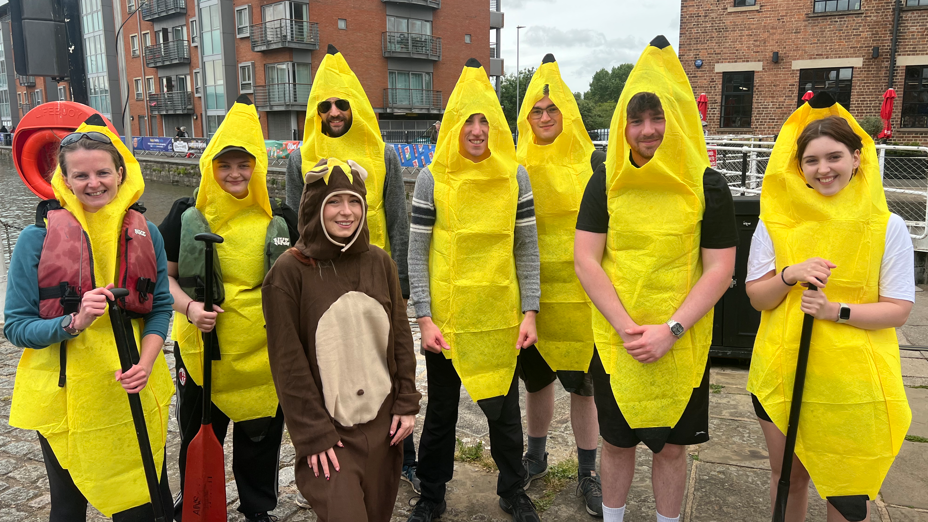 Seven people are wearing banana costumes standing in a row, and a single person is wearing a bear costume. They are standing and looking at the camera smiling.