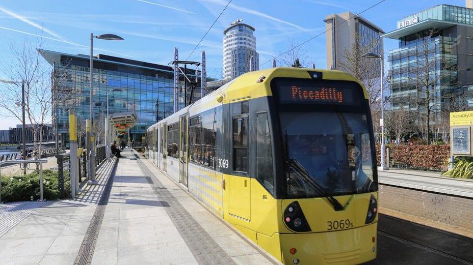 A Metrolink tram with its yellow livery by the platform at the BBC's northern headquarters in MediaCity, Salford.