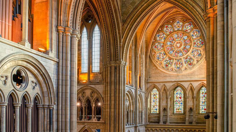 The interior of Truro Cathedral is lit up with light shining through a multi-coloured circular stained glass rose window. There are also perpendicular columns in the gothic revival style the cathedral is built in.