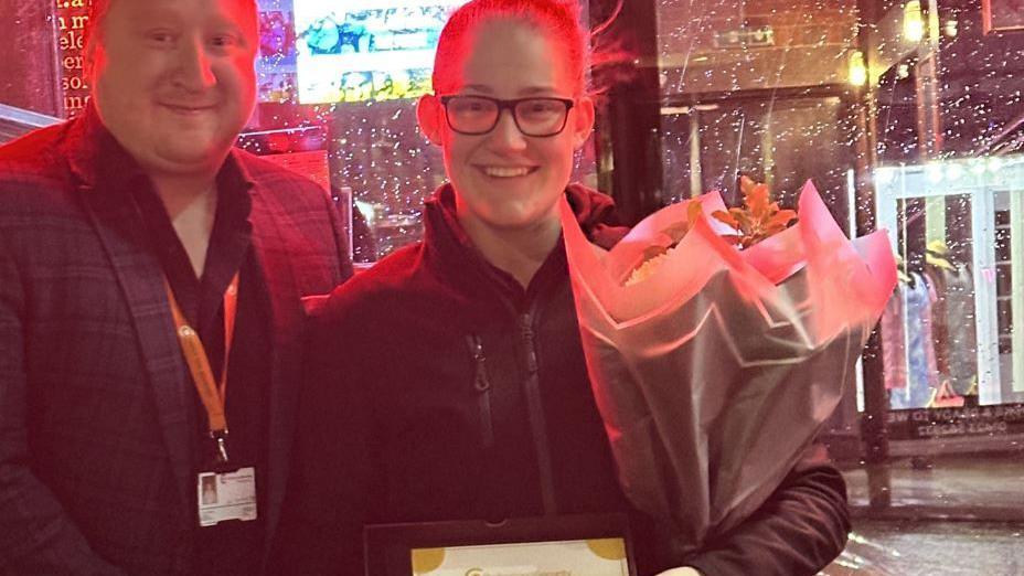 Woman wearing glassses holding a bunch of flowers in the centre stood with a man wearing a a dark jacket and shirt