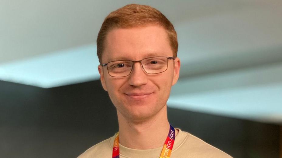 Ross Greer, a man with short ginger hair and glasses, smiles at the camera. He wears a rainbow lanyard and a beige jumper