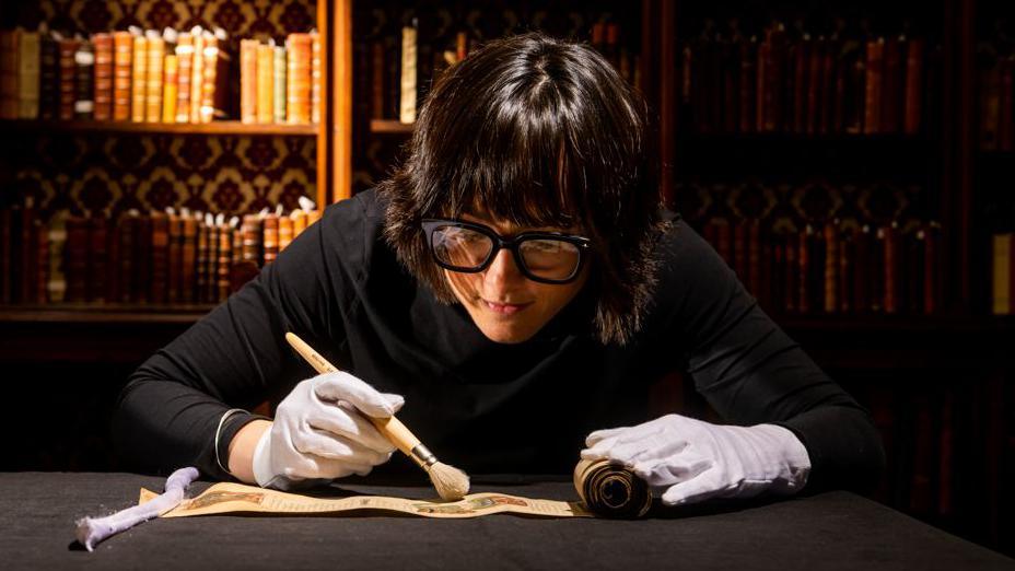 A woman with a black bob and fringe wearing large black-rimmed glasses sits in a library brushing a medieval manuscript with a brush. She is wearing a black polo neck and white gloves.